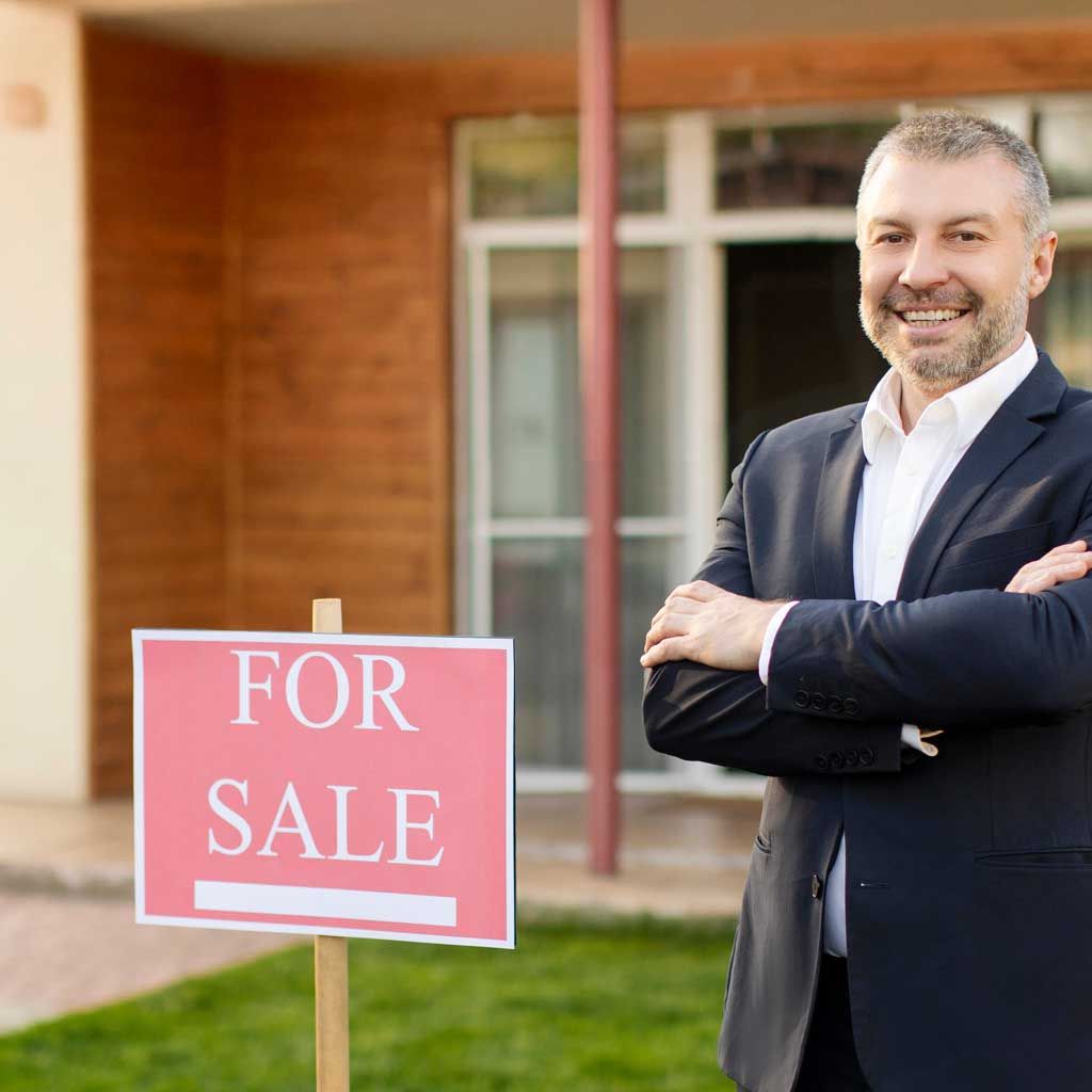 A business owner in a suit stands with his arms crossed outside a property, beside a for-sale sign.