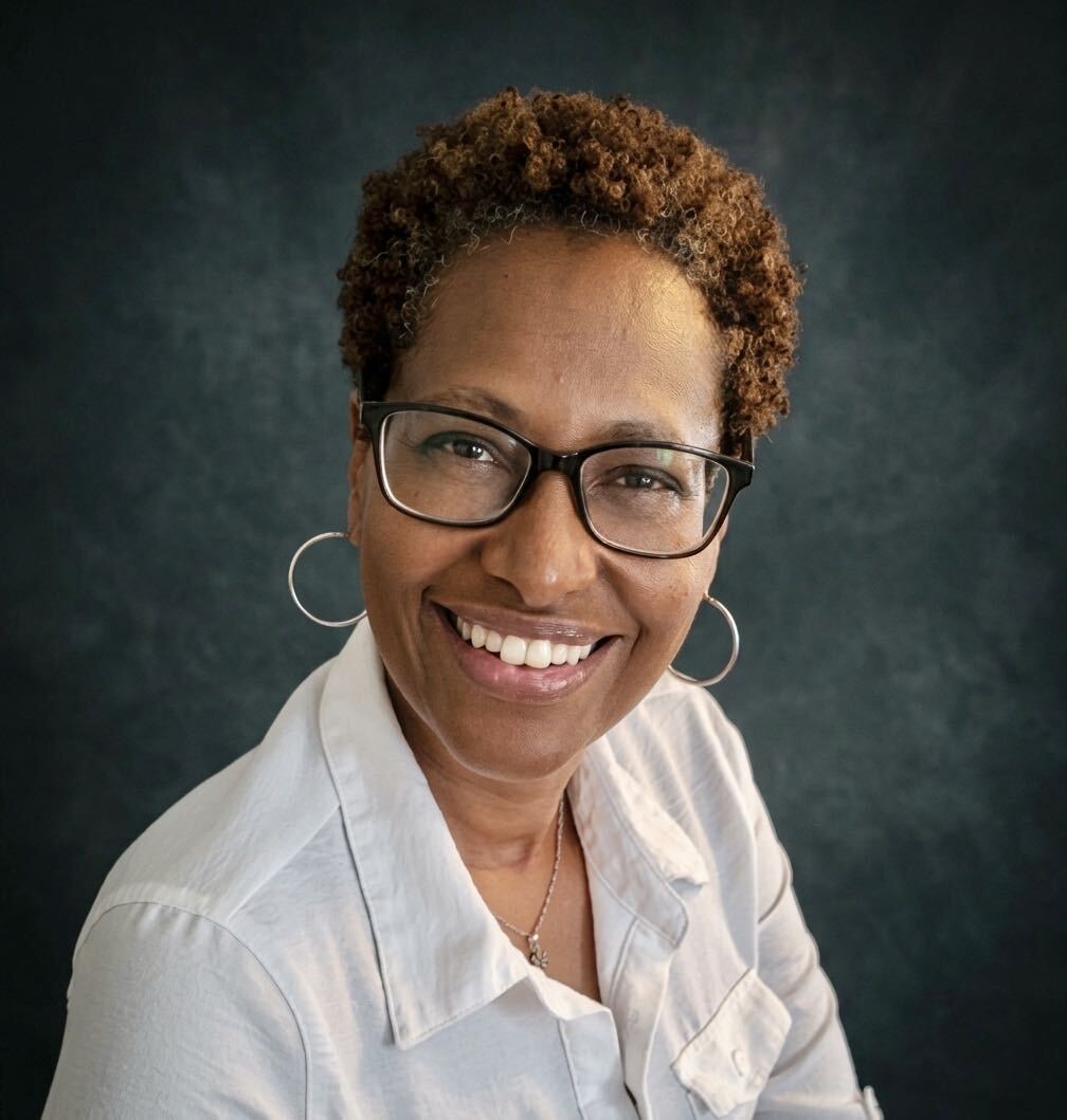 Beth Sweatman, wearing glasses and a white shirt, smiling against a dark background.