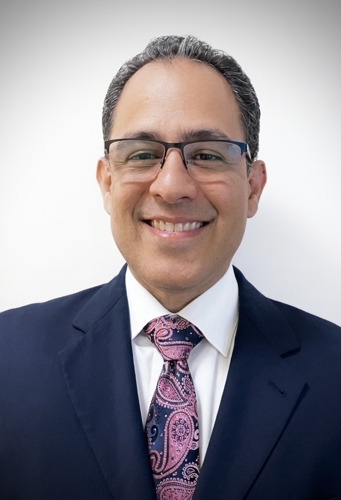 Dan Barrientos, in glasses, a navy suit and a patterned tie, smiling against a white background.