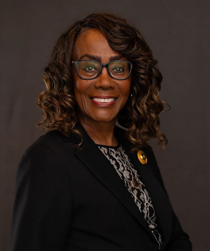 Dianne Oliphant, with dark curly hair and glasses, in a black jacket against a dark background.