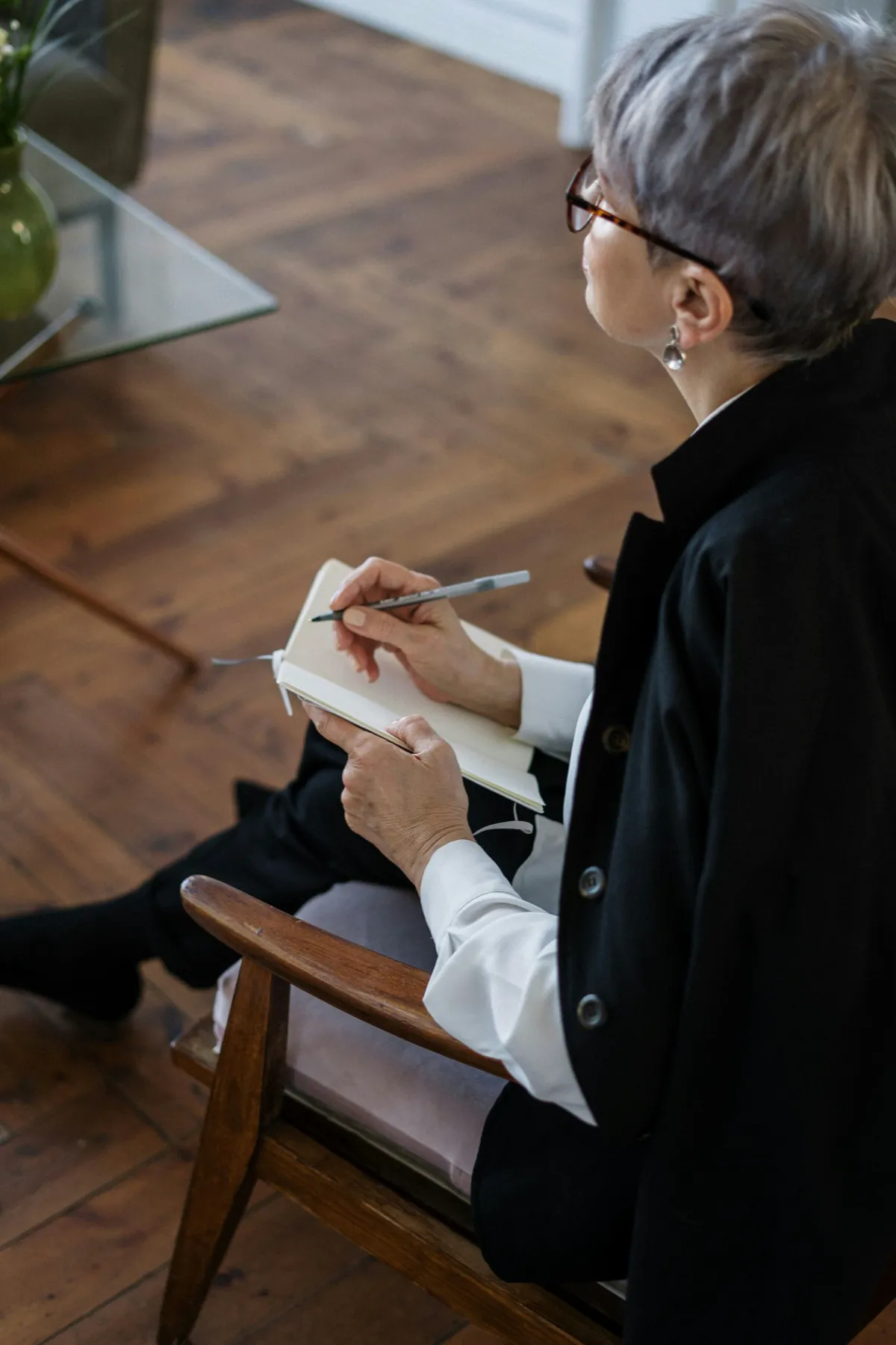 A person in a black blazer sits in a wooden chair, taking notes in a small journal.